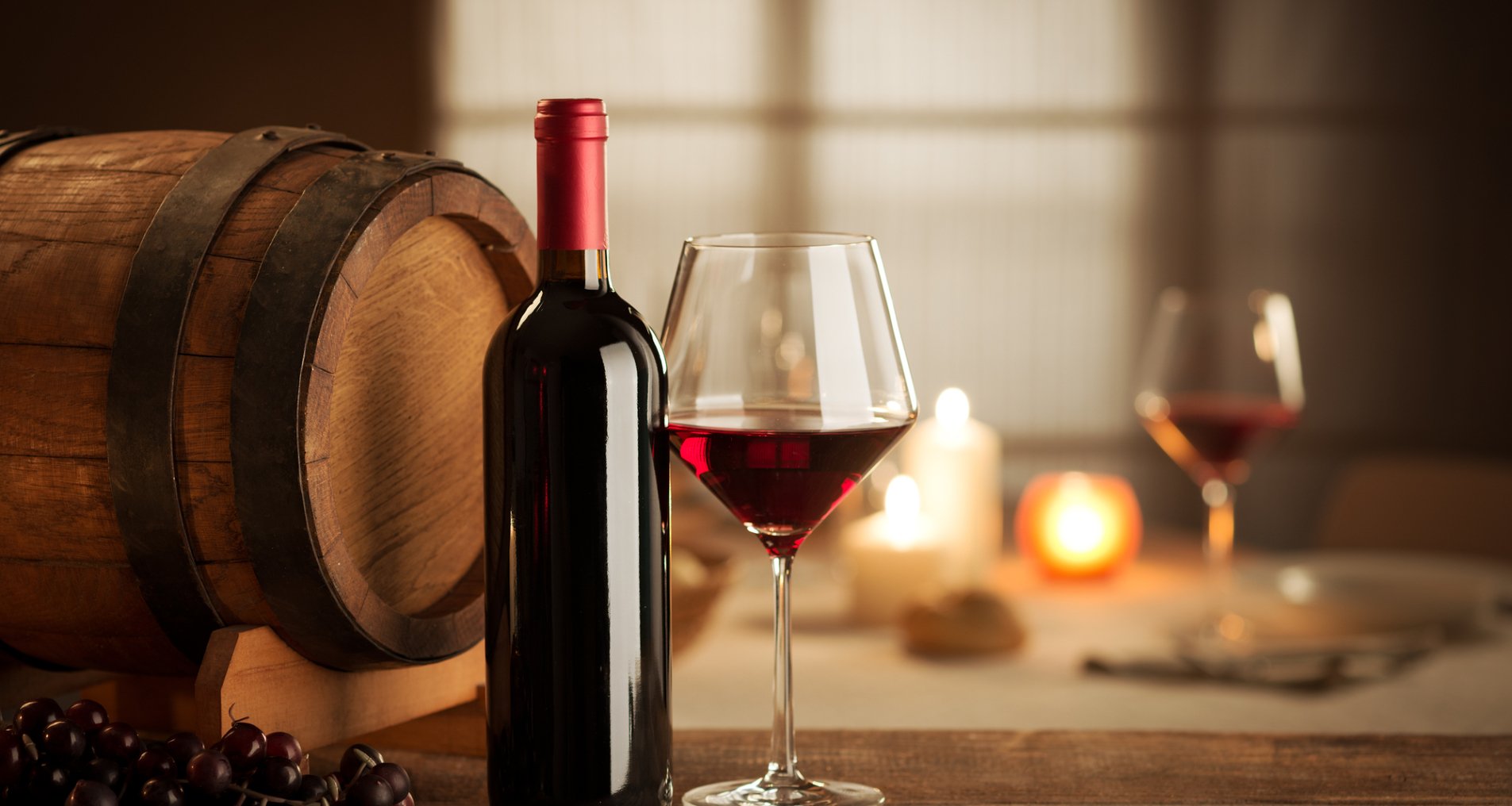 Пляшка для вина чорного кольору та келих з налитим червоним вином на фоні бочки
