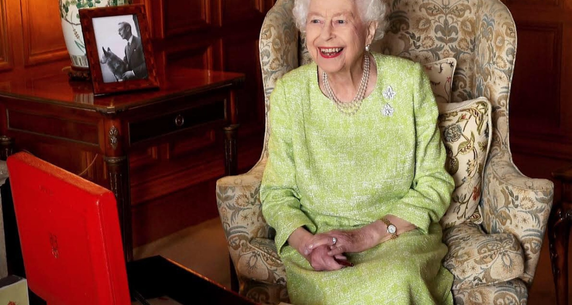 Королева Елизавета в зеленом костюме сидит в кресле напротив красного кейса