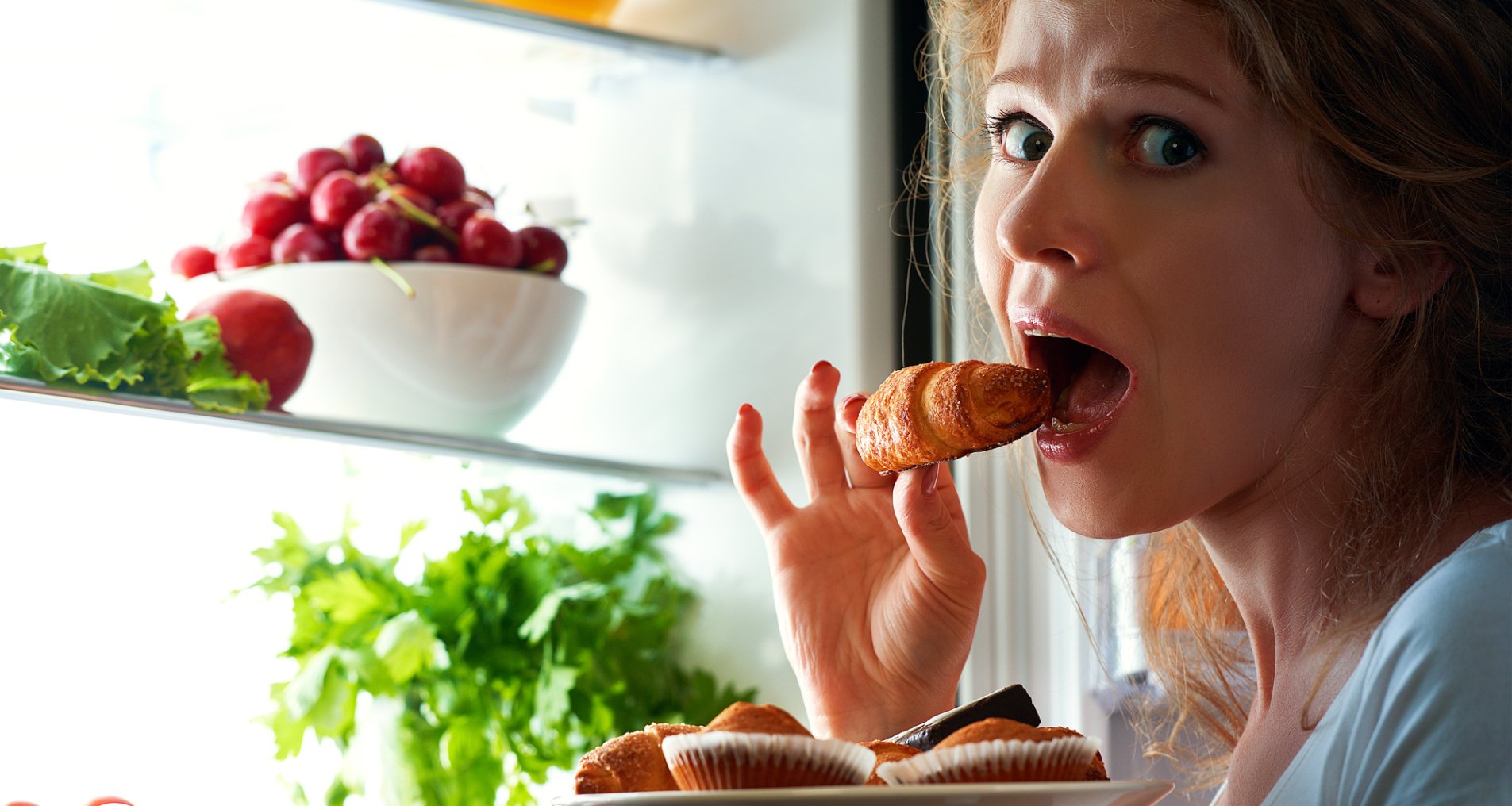 Женщина ест круассан возле холодильника.
