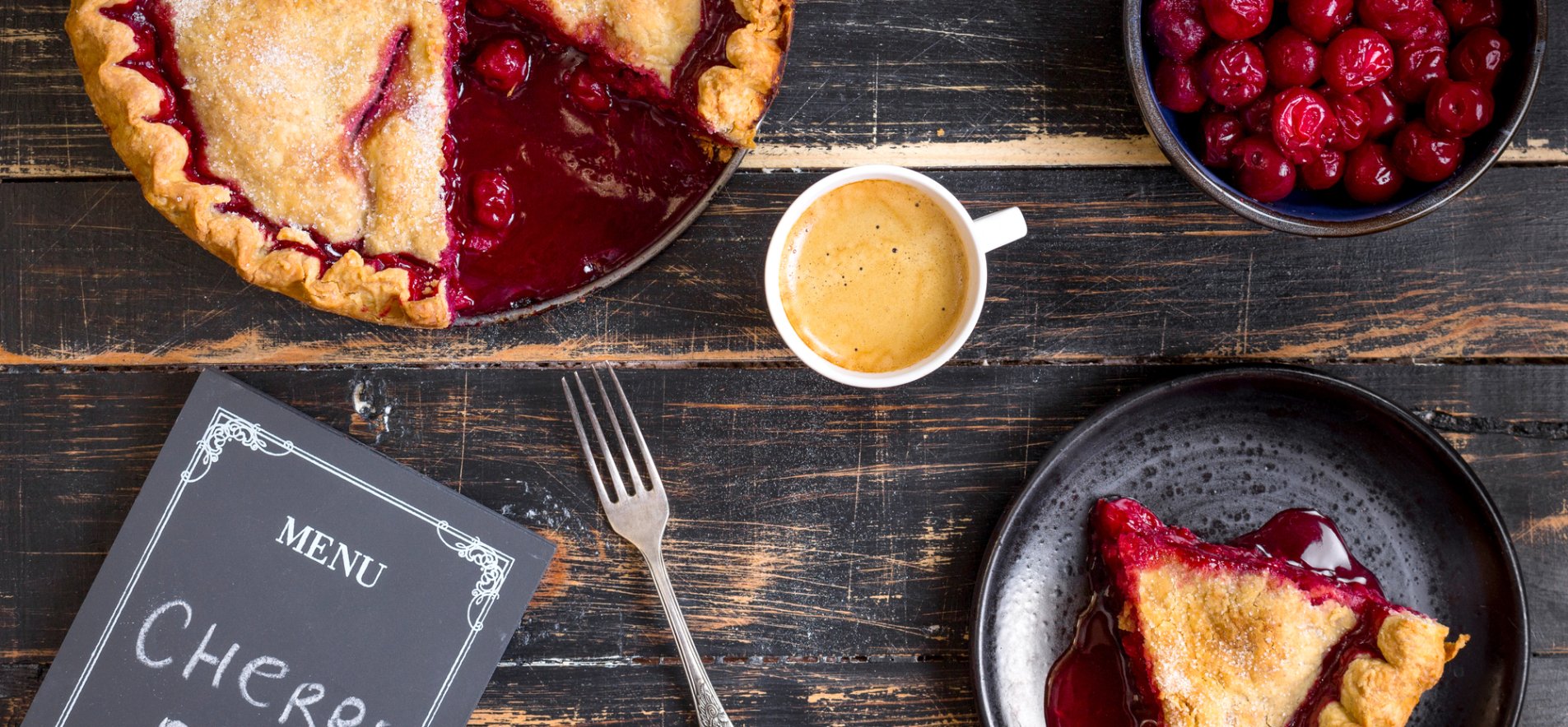 Вишневый пирог, тарелка с вишнями, кусок вишневого пирога и чашка кофе на деревянном столе.